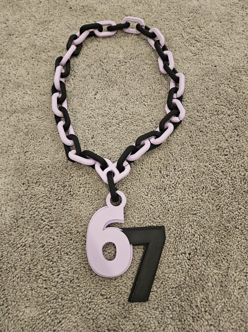 May include: A chunky necklace featuring a chain design with alternating black and light purple links. A light purple pendant with the number 6 and a black number 7 hangs from the chain.