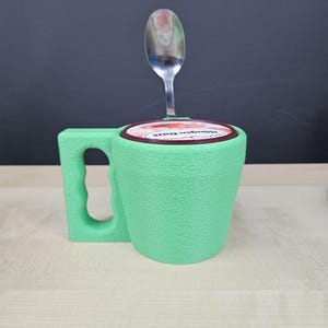 Pu&ograve; includere: Una tazza verde chiaro con una superficie strutturata, un manico e un coperchio con un'etichetta rossa e bianca. Un cucchiaio d'argento &egrave; inserito nel coperchio. La tazza sembra contenere un contenitore di gelato.