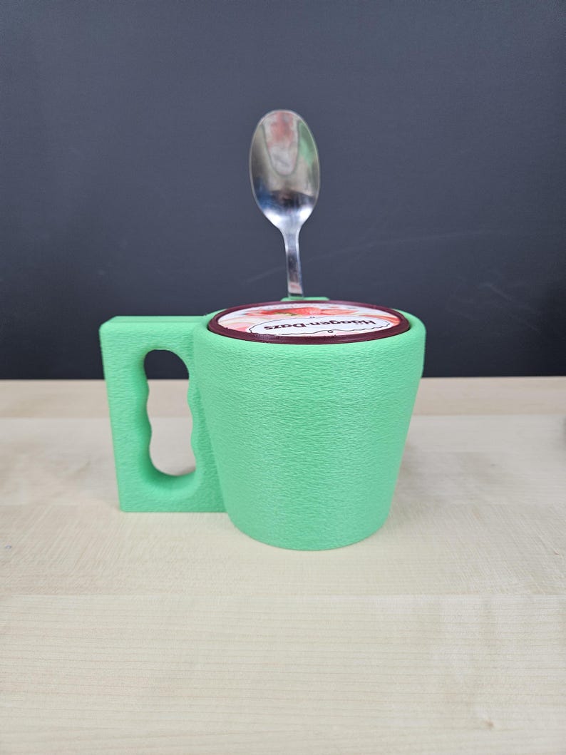 Pu&ograve; includere: Una tazza verde chiaro con una superficie strutturata, un manico e un coperchio con un'etichetta rossa e bianca. Un cucchiaio d'argento &egrave; inserito nel coperchio. La tazza sembra contenere un contenitore di gelato.
