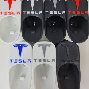 May include: Six 3D printed Tesla key holders in various colors, including white, black, and gray. Each holder features the Tesla logo and the word "TESLA" printed on the front.