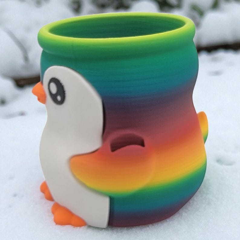 May include: A colorful penguin-shaped container with a rainbow gradient design. The penguin has a white face, black eyes, and orange beak and feet. The container is likely a decorative item or a small planter.