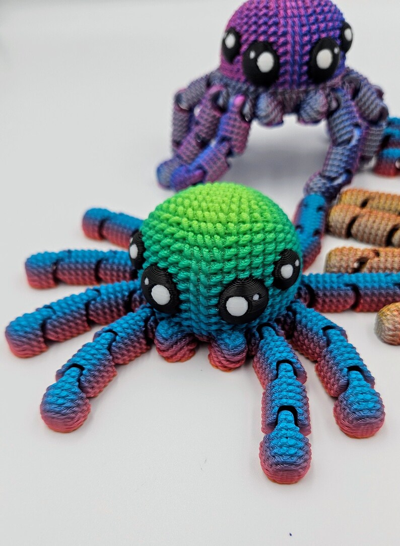 Op de afbeelding: Twee kleurrijke 3D-geprinte octopus speelgoedjes. E&eacute;n is blauw, groen en roze, de andere is paars en blauw. Beide hebben grote zwarte ogen.
