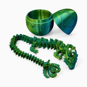 May include: A green and blue 3D printed dragon figurine with a segmented tail. The dragon is posed in a curled position with its head raised. The dragon is inside a green and blue egg-shaped container with a heart design.