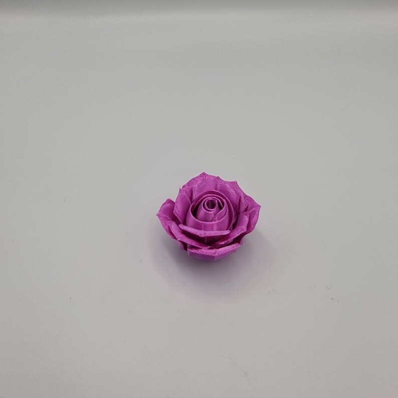 May include: A single, vibrant purple rose. The rose is in full bloom, with tightly packed petals forming a spiral center. The petals have a textured appearance, and the overall shape is round. The rose is set against a plain, light gray background.
