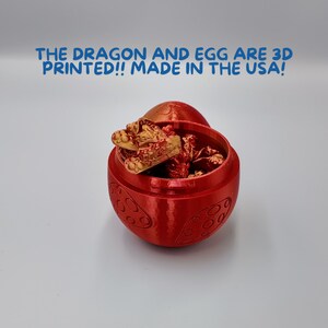 May include: A red 3D printed egg with a golden dragon figurine inside. The egg has a textured surface with a repeating pattern. The text "THE DRAGON AND EGG ARE 3D PRINTED!! MADE IN THE USA!" is displayed above the egg.