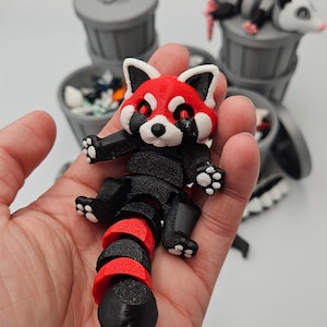 May include: A 3D-printed red panda figurine with a black and red segmented body, held in a hand. The panda has a red face with white accents and black paws. Other figurines are in the background.