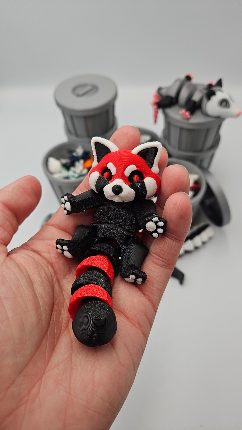 May include: A 3D-printed red panda figurine with a black and red segmented body, held in a hand. The panda has a red face with white accents and black paws. Other figurines are in the background.