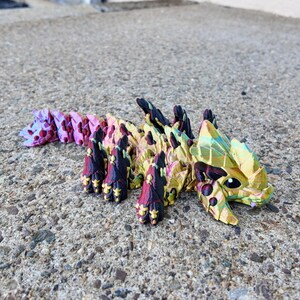 May include: A colorful origami dragon made of paper with a gold, green, pink, and black color scheme. The dragon is lying on a gray concrete surface.