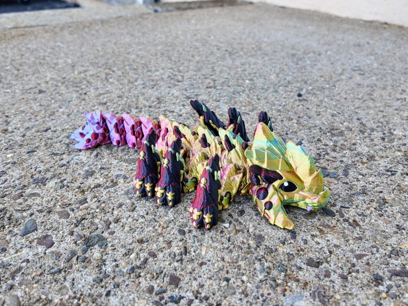 May include: A colorful origami dragon made of paper with a gold, green, pink, and black color scheme. The dragon is lying on a gray concrete surface.