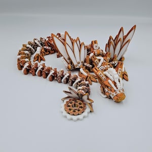 May include: A white and copper-coloured 3D printed dragon figurine with intricate details. The dragon has wings, a long tail, and a gear-like design. The figurine is on a white background.