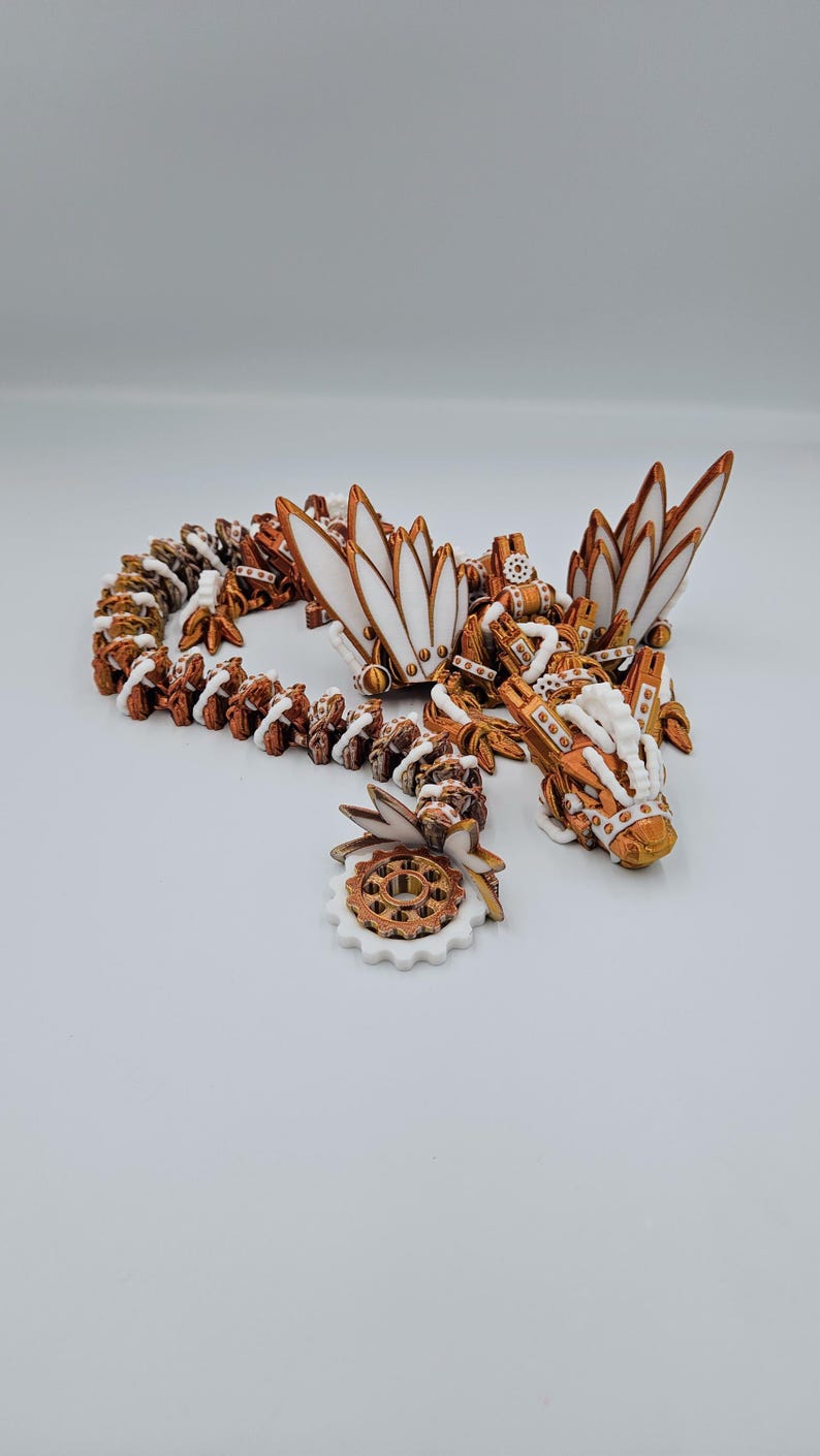 May include: A white and copper-coloured 3D printed dragon figurine with intricate details. The dragon has wings, a long tail, and a gear-like design. The figurine is on a white background.