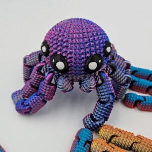 Op de afbeelding: Een 3D-geprinte octopus met een paarse en blauwe iriserende afwerking. De octopus heeft grote, witte ogen en acht gelede poten. De octopus zit op een wit oppervlak.
