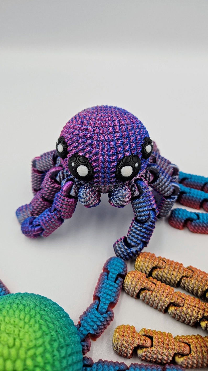 Op de afbeelding: Een 3D-geprinte octopus met een paarse en blauwe iriserende afwerking. De octopus heeft grote, witte ogen en acht gelede poten. De octopus zit op een wit oppervlak.