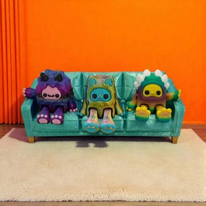 Puede incluir: Tres figuras de dibujos animados coloridas sentadas en un sof&aacute; turquesa. Las figuras tienen ojos grandes y dise&ntilde;os &uacute;nicos, incluyendo cuernos y rasgos animales. El sof&aacute; est&aacute; colocado sobre una alfombra esponjosa de color crema, con un fondo naranja.