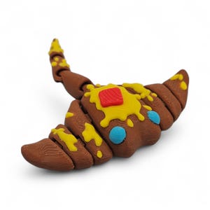 May include: A brown, croissant-shaped sculpture with yellow and red accents, and blue dots. The pastry-like object has a textured surface and is set against a white background. The sculpture is a whimsical, food-themed art piece.