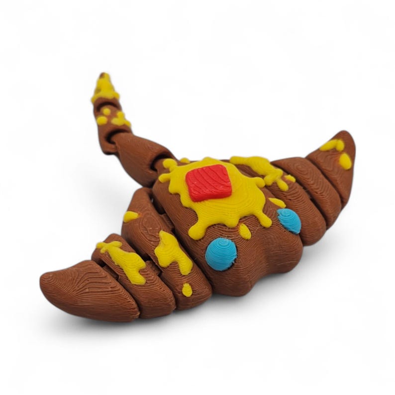 May include: A brown, croissant-shaped sculpture with yellow and red accents, and blue dots. The pastry-like object has a textured surface and is set against a white background. The sculpture is a whimsical, food-themed art piece.