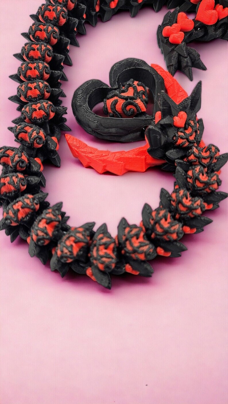 Op de afbeelding: Een zwarte en rode ketting met een hartvormige hanger. De hanger is gemaakt van zwart plastic en heeft een rood hartontwerp. De ketting is gemaakt van zwarte plastic kralen met rode accenten.