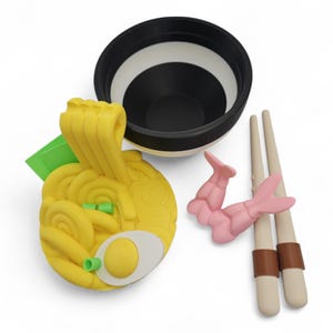 K&ouml;nnte beinhalten: Ein skurriles Ramen-Sch&uuml;ssel-Set mit einer schwarz-wei&szlig; gestreiften Sch&uuml;ssel, gelben Nudeln mit Ei, gr&uuml;ner Garnitur, rosa Garnelen und beigefarbenen Essst&auml;bchen mit braunen B&auml;ndern. Ein lustiger, verspielter Artikel zum Thema Essen.