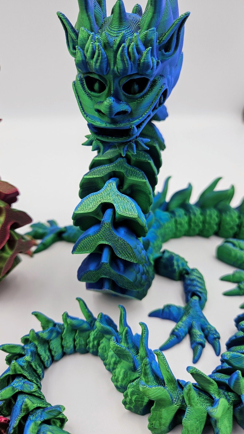 May include: A 3D-printed articulated dragon in shades of blue and green. The dragon has a detailed head, a segmented body, and clawed feet. The color gradient gives the dragon a textured appearance, and it is posed on a white surface.