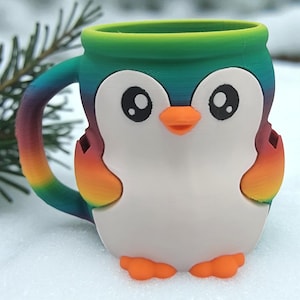 May include: A colorful penguin-shaped mug with a rainbow handle and accents. The mug features a white body, black eyes, and an orange beak and feet. The rim of the mug is green. The mug is sitting on a snowy surface.