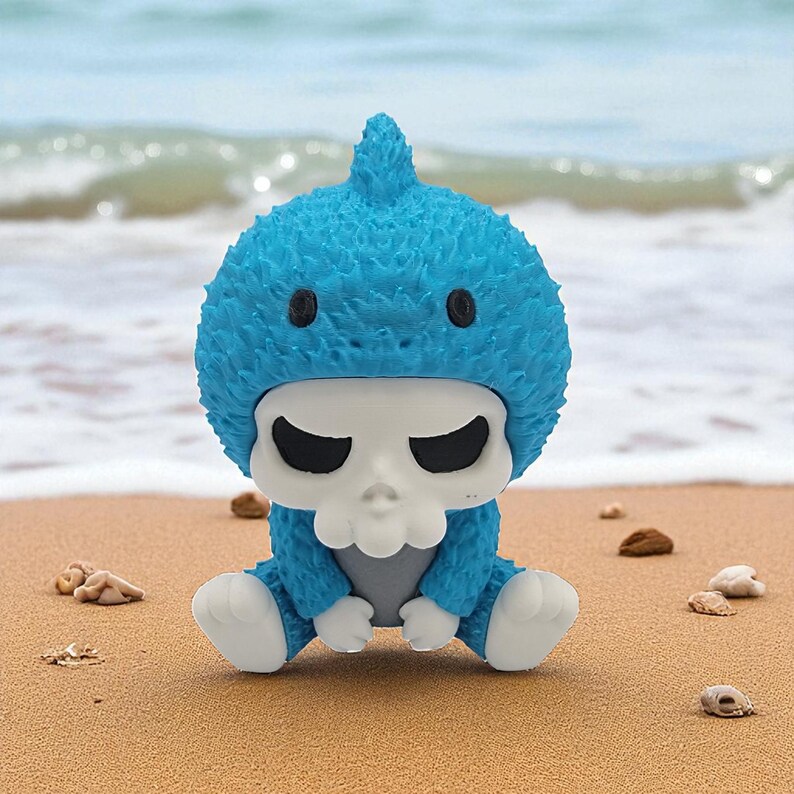 May include: A blue figurine of a baby-like creature with a skull face and a textured, spiky head covering. The figure is sitting on a sandy beach with the ocean in the background. The figure has black eyes and white hands and feet.