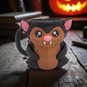 May include: A Halloween-themed mug shaped like a cartoon bat. The mug features a black exterior with a brown belly, pink inner ears, and a pink nose. The bat has large, round eyes and small fangs. The mug is sitting on a wooden surface.