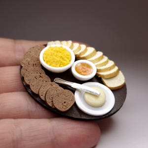 Puede incluir: Arreglo de comida en miniatura sobre una fuente ovalada marrón oscuro. Incluye pan en rodajas, rebanadas de pan marrón, dos cuencos blancos pequeños con contenido amarillo y naranja, y un plato pequeño con mantequilla y un cuchillo. Una exhibición de comida detallada.