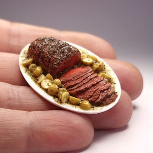 Puede incluir: Un plato ovalado en miniatura que presenta un asado de carne roja cocinado y rebanado, sazonado con hierbas oscuras, acompañado de pequeñas verduras redondas y claras en una salsa ligera. El plato se presenta sobre una bandeja blanca y lisa.