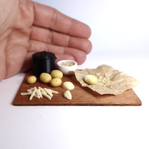 May include: Miniature kitchen scene with a black pot, small white bowl, potatoes, garlic cloves, and fries on a wooden cutting board. A piece of parchment paper holds a peeled potato. The scene is set against a white background.