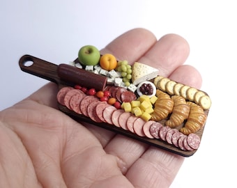 Puede incluir: Una tabla de embutidos en miniatura con una variedad de alimentos. Incluye salami, queso, uvas y cruasanes. La tabla de madera marrón oscuro tiene un asa. Los alimentos son de varios colores, incluyendo rojo, verde y amarillo.