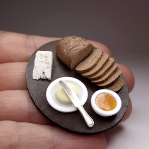 Puede incluir: Arreglo de comida en miniatura sobre una bandeja marrón oscuro. Incluye pan en rodajas, una cuña de queso, una porción de mantequilla con un cuchillo y un cuenco pequeño de mermelada. El pan es marrón y la mermelada es dorada.