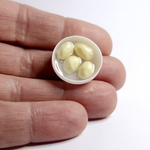 Op de afbeelding: Miniatuur witte kom gevuld met vier lichtgele, ronde objecten in een heldere vloeistof. De kom wordt in de hand van een persoon gehouden. De objecten lijken voedsel te zijn.