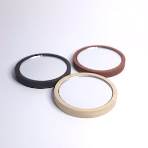 May include: Three small, round mirrors with wooden frames in black, brown, and natural wood tones. The mirrors are arranged on a white surface, showcasing their reflective surfaces and simple design. Each mirror is approximately 7.6 cm in diameter.
