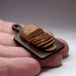 Puede incluir: Miniatura de pan marrón, parcialmente rebanado, sobre una pequeña tabla de cortar de madera marrón oscuro. El pan tiene una superficie texturizada y está cortado en varias rebanadas. La tabla de cortar tiene un asa con un agujero.