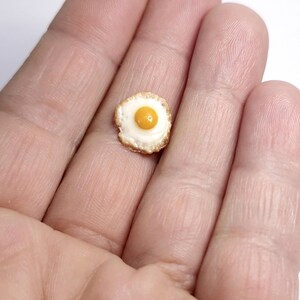 May include: A miniature fried egg with a yellow yolk and white egg white, with a golden-brown edge. The tiny food item is held in a person's hand, showcasing its small size. This could be a charm, a dollhouse accessory, or a piece of miniature food art.