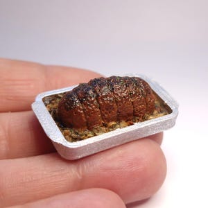Op de afbeelding: Miniatuur rosbief in een zilveren bakvorm. De braadstuk is donkerbruin met een textuur en ligt op een bedje van kruiden en groenten. Een gedetailleerd voedselitem voor poppenhuizen of miniatuurcollecties.