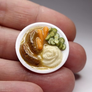 Peut inclure: Une assiette miniature de nourriture comprenant deux saucisses, des carottes, des petits pois et de la purée de pommes de terre. La nourriture est disposée sur une assiette blanche. L'image est tenue entre deux doigts.