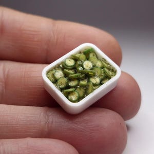 Puede incluir: Pequeño plato rectangular blanco lleno de pepinos verdes en rodajas en miniatura. Los pepinos están sazonados con pimienta negra y hierbas. Esta réplica a pequeña escala es probablemente para una casa de muñecas o una colección en miniatura.