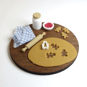 May include: Miniature baking scene with a wooden rolling pin, gingerbread man cookie cutter, and a round wooden board. Includes a small white jar, a bowl of red jam, a blue and white checkered napkin, and gingerbread man biscuits.