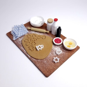 May include: A miniature baking scene on a wooden board. It includes rolled-out dough with cookie cutouts, flour, a rolling pin, small bowls with ingredients, and bottles. A blue and white checkered cloth is also present.