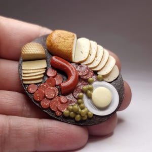 Puede incluir: Miniatura de tabla de embutidos con salami, queso, pan, aceitunas y una salchicha. La comida está dispuesta sobre una fuente ovalada oscura. Los alimentos en miniatura son detallados y realistas.