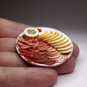 Puede incluir: Una tabla de embutidos en miniatura con carnes en rodajas, queso y un pequeño cuenco con salsa. La tabla presenta varias tonalidades de carnes rojas y marrones, rebanadas de queso amarillo y bayas rojas y amarillas. Un cuenco blanco diminuto contiene una salsa verde.