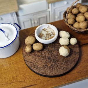 Może przedstawiać: Miniaturowa scena kuchenna z białym emaliowanym garnkiem z niebieskim wykończeniem, drewnianą deską do krojenia i małą białą miską wypełnioną jedzeniem. Kilka całych i obranych ziemniaków ułożonych jest na desce i w wiklinowym koszu.