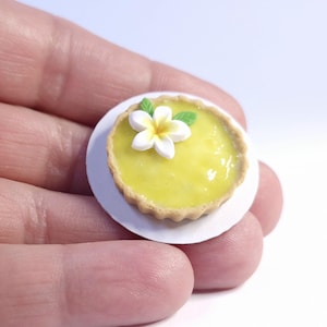 Puede incluir: Mini tarta de limón sobre un plato blanco, cubierta con una flor blanca y amarilla y hojas verdes. La tarta tiene un relleno amarillo y una corteza dorada. Pequeño postre.