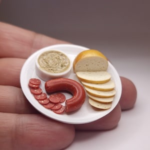 Puede incluir: Plato de comida en miniatura con salchicha roja, pan en rodajas, mostaza y rodajas de salchicha. El pan es de color marrón claro y la mostaza está en un cuenco blanco. El plato es blanco.