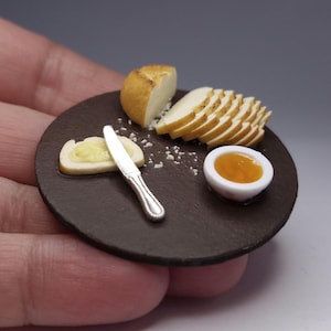 Puede incluir: Mini tabla de embutidos con pan, queso, mantequilla y mermelada. La tabla redonda, de color marrón oscuro, contiene una rebanada de pan con mantequilla, un cuchillo plateado, una cuña de queso, pan en rodajas y un cuenco pequeño de mermelada.