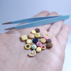 May include: A collection of miniature, colorful candies and sweets, including cookies, buttons, and a lemon slice, are held in a hand. A pair of light blue tweezers is positioned above the treats.