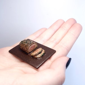 May include: Miniature cooked steak with a slice, presented on a small brown rectangular plate. The steak is seasoned with herbs and spices. The miniature food item is held in a person's hand.