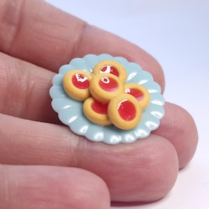 Puede incluir: Un plato en miniatura de color azul claro con borde festoneado, que contiene seis galletas pequeñas y redondas. Cada galleta tiene un centro de mermelada roja y una base de galleta de color amarillo-marrón. Las galletas están dispuestas en una pequeña pila en el plato.