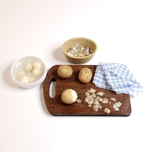 Op de afbeelding: Een miniatuurkeuken met een houten snijplank, aardappelen en gehakte uien. Een kleine kom met geschilde aardappelen en een gevlochten mand met gehakte uien zijn ook aanwezig. Een blauw-wit geruite doek.